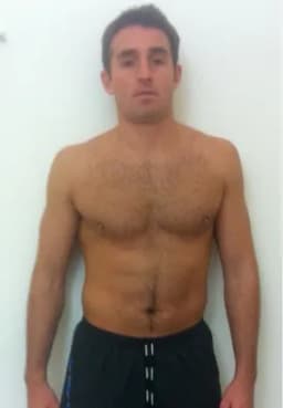 Shirtless man with short dark hair standing against a white background, wearing dark shorts.