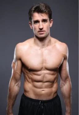 Muscular shirtless man with defined abs posing against a dark background, wearing black shorts.